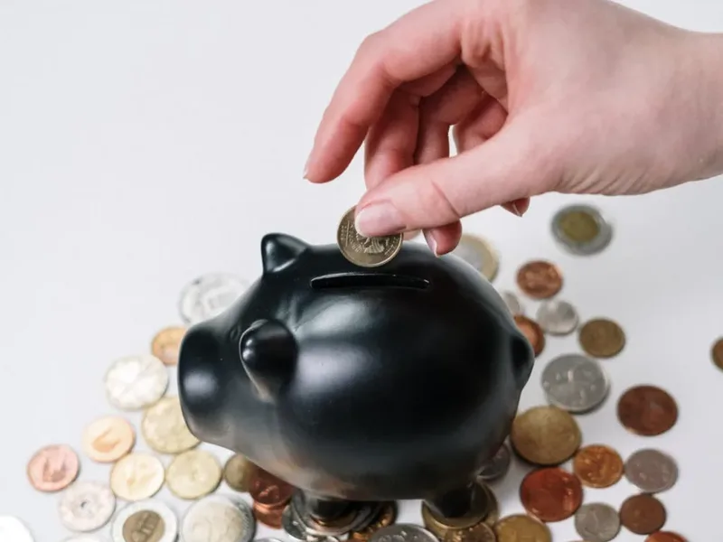 Une main glisse une pièce dans une tirelire noire, entourée de monnaie
